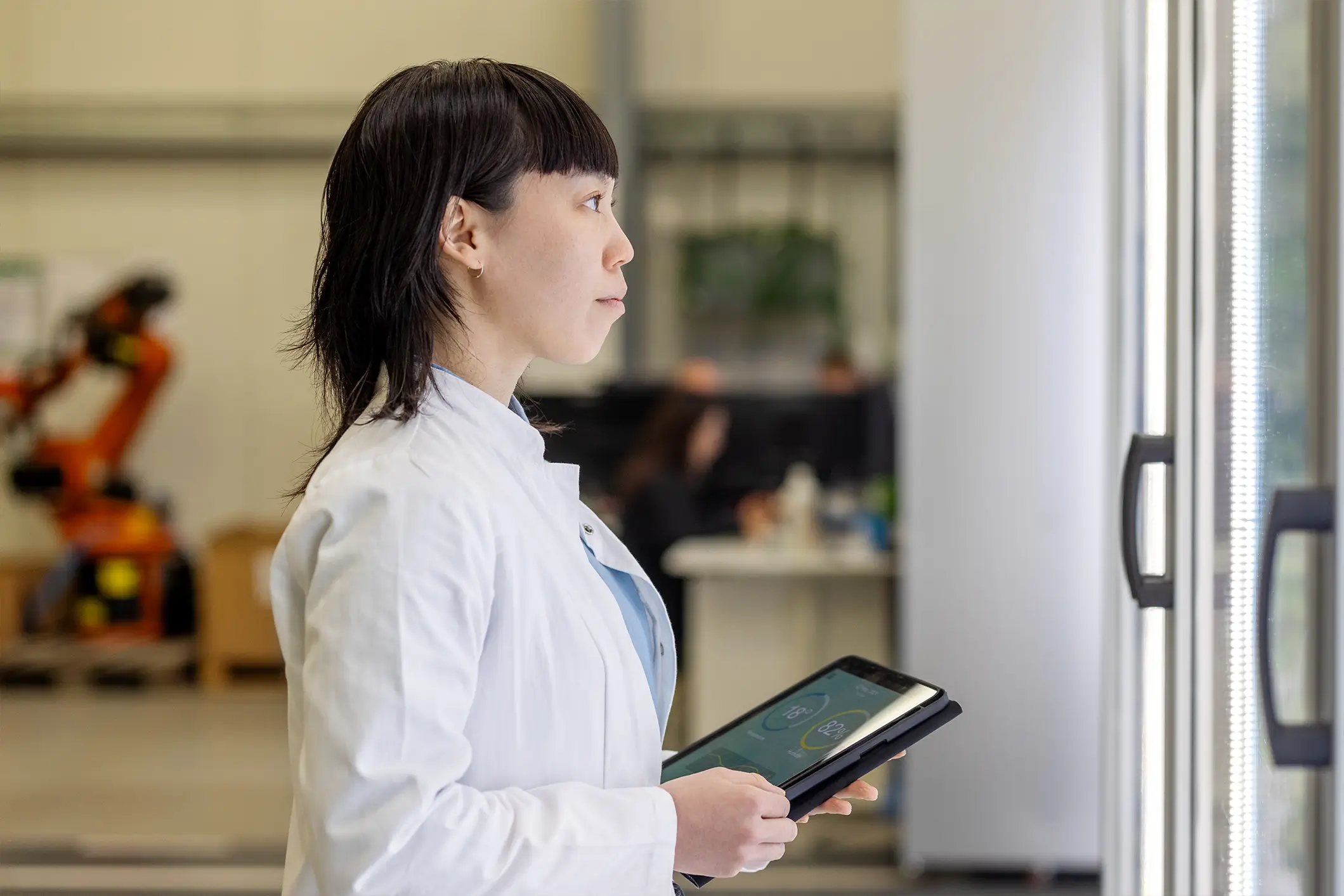 A woman in a white lab coat holds a tablet, standing indoors near glass doors. In the background, a robotic arm and people working are slightly visible, suggesting a laboratory or industrial setting.