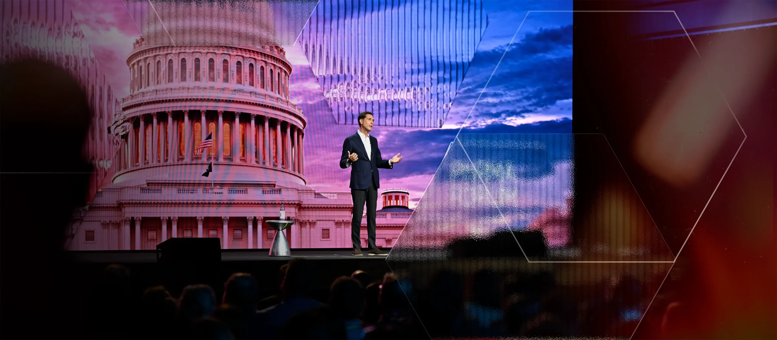 A person stands on a stage, speaking in front of a large backdrop featuring an image of the U.S. Capitol building against a vibrant sky. Audience members are visible in silhouette in the foreground.