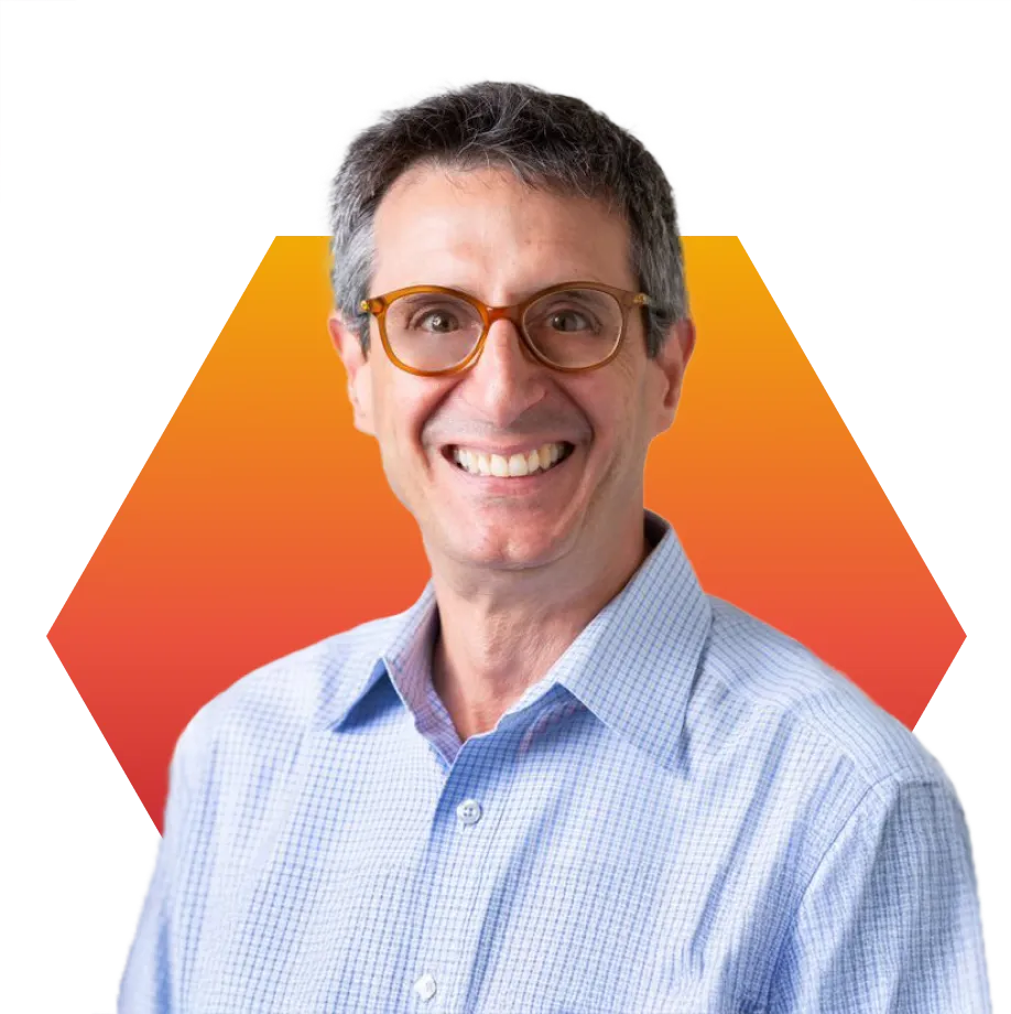 A smiling man with short gray hair and glasses, wearing a light blue checkered shirt, stands in front of an orange hexagon background.