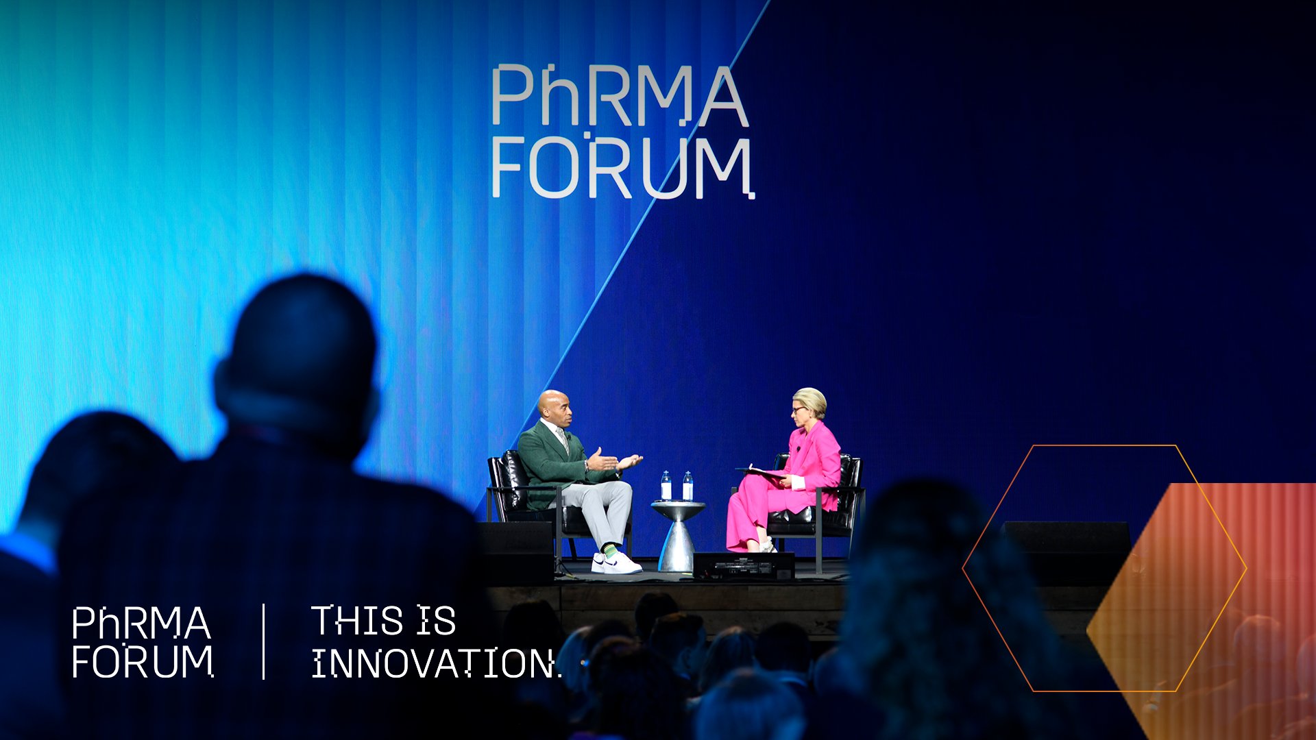 Two people sit onstage in conversation at the PhRMA Forum, with a large audience watching. The stage background is blue with the words "PhRMA FORUM" and "THIS IS INNOVATION." displayed.