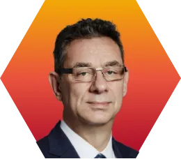 A man with short dark hair and glasses, wearing a dark suit and tie, is shown against an orange-red geometric background.