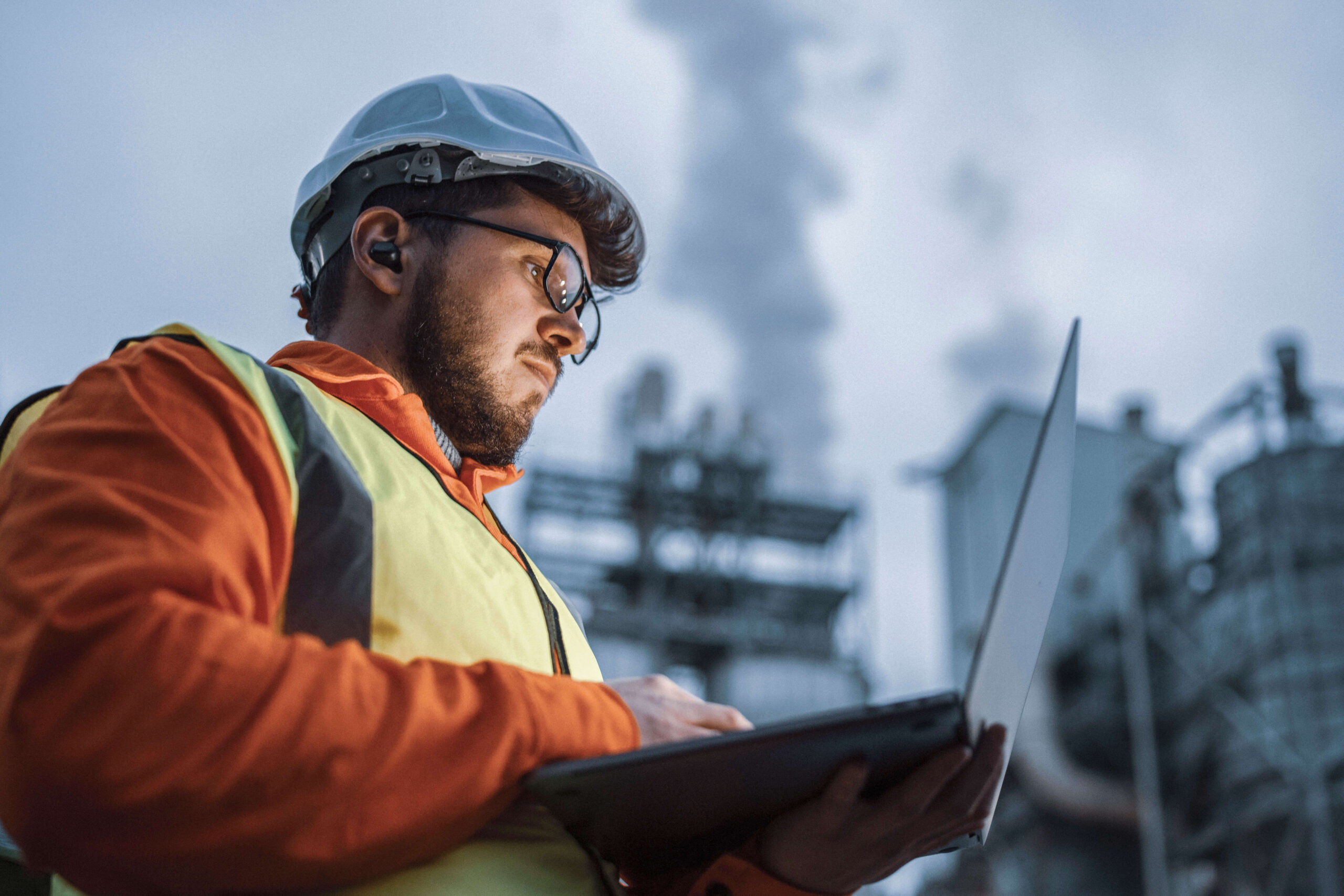 A worker wearing a hard hat, safety glasses, and a reflective vest uses a laptop outdoors at an industrial site with machinery and smoke in the background.