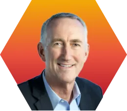 A smiling middle-aged man with short gray hair, wearing a dark suit jacket and light blue shirt, is set against an orange and yellow gradient hexagonal background.