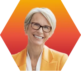 Smiling woman with short gray hair and glasses, wearing a yellow blazer and a white top, standing in front of an orange-red gradient hexagonal background.
