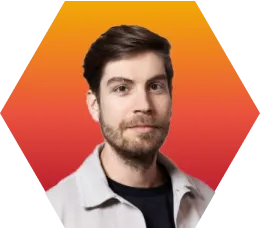 A man with short brown hair and a beard, wearing a light-colored jacket over a dark shirt, stands in front of a hexagonal orange-to-red gradient background.