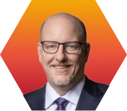 A smiling man with glasses, a goatee, and a bald head wears a dark suit, light shirt, and purple tie, posed in front of an orange-to-red gradient background.