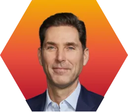 A man with short brown hair, wearing a navy blazer and light blue shirt, is smiling slightly in front of a red and orange hexagonal gradient background.