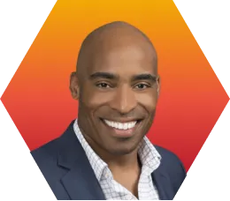 A smiling man with a shaved head in a blue blazer and open-collared shirt is shown in front of an orange and yellow gradient geometric background.