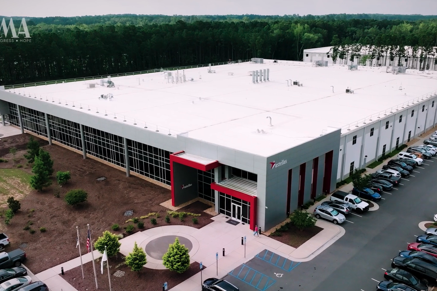 Aerial view of a large, modern industrial or commercial building with a white roof, gray and red exterior, surrounded by parked cars, greenery, and trees in the background.