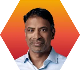 A man with short dark hair and some gray, wearing a light blue collared shirt, is shown against an orange and red hexagonal gradient background.