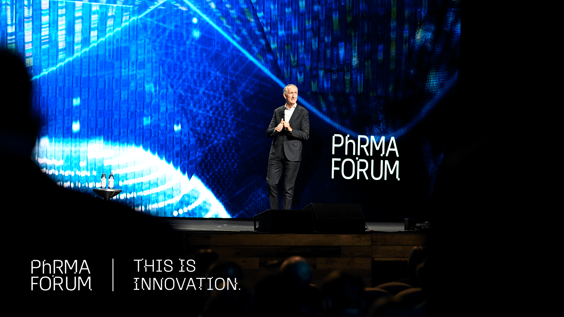 A man in a suit stands on stage speaking at the PhRMA Forum, with a large blue digital background behind him and the words "PhRMA Forum" and "This Is Innovation" displayed on the image.