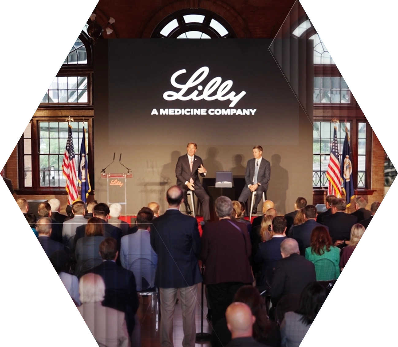 A large group of people watches two speakers on stage at a Lilly medicine company event, with flags, a podium, and a large screen displaying the company logo in the background.