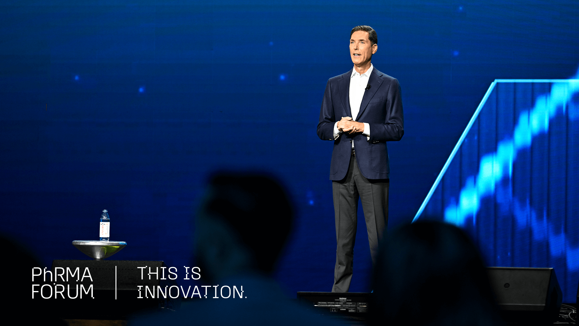 A man in a suit stands on a stage giving a presentation. A water bottle sits on a small table nearby. The background is blue and displays the text "PhRMA Forum | This is innovation.