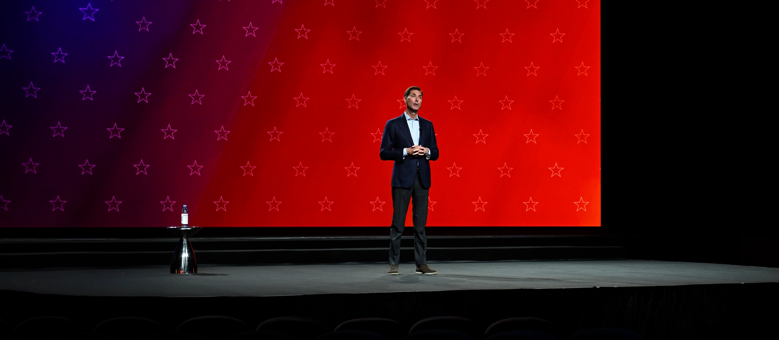 A man in a suit stands on a stage with a red background decorated with stars. A small round table with a water bottle is to his left. The setting suggests a formal presentation or speech.