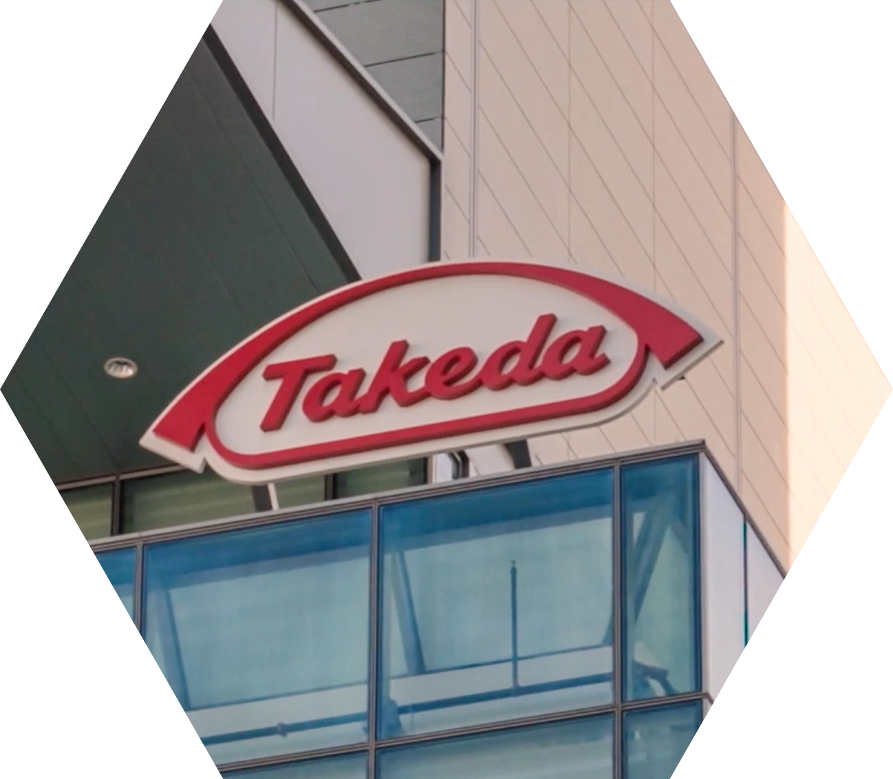 The image shows the Takeda logo on the exterior of a modern glass building. The logo features the word "Takeda" in red cursive font inside a red arch shape.