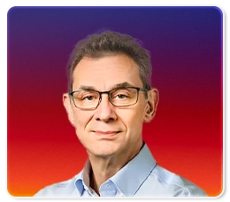 A middle-aged man with short gray hair and glasses, wearing a light blue collared shirt, stands against a gradient background of purple, red, and orange.