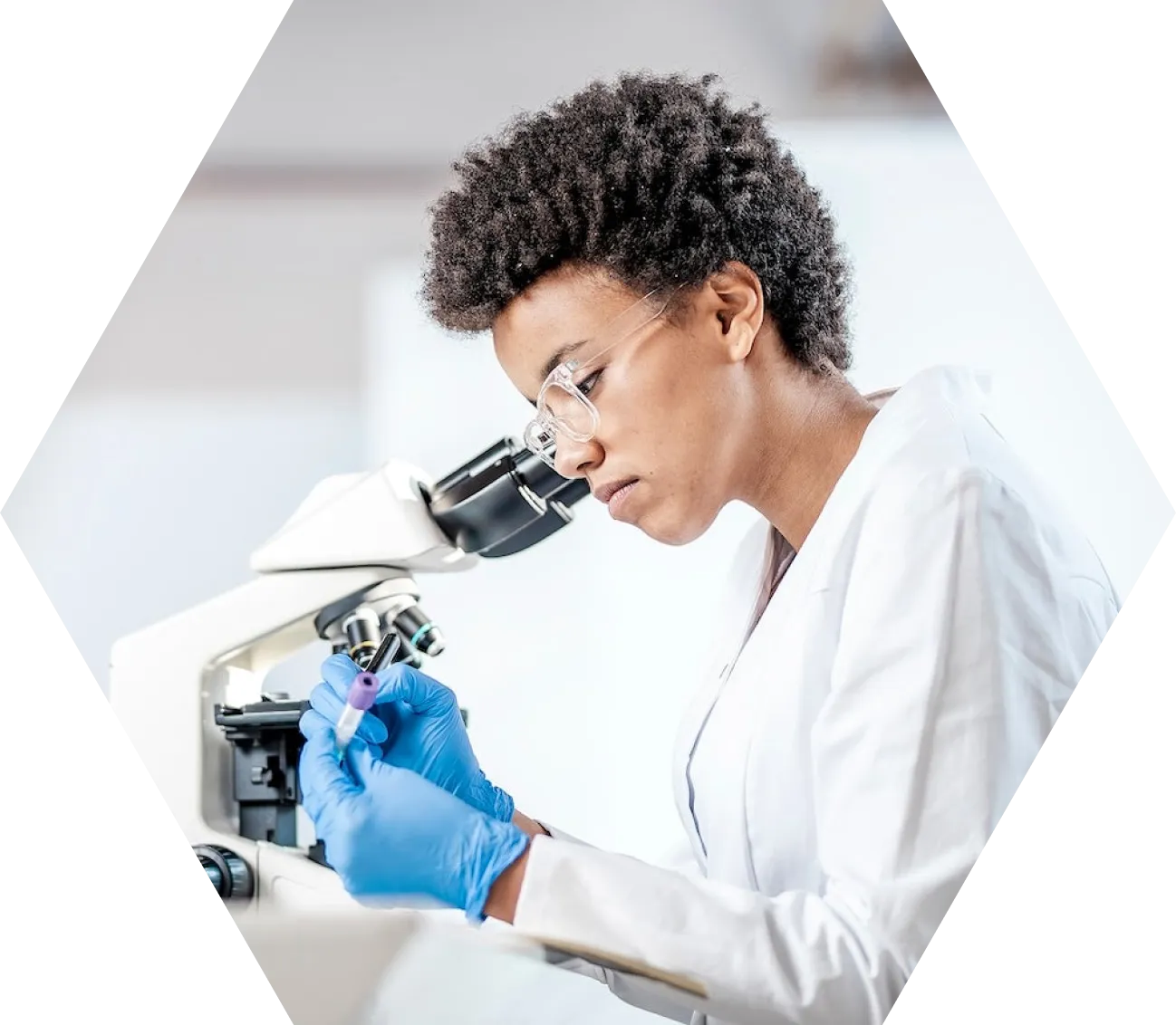 A scientist wearing glasses and blue gloves examines a sample in a test tube while sitting at a microscope, focused on her research in a bright laboratory setting.